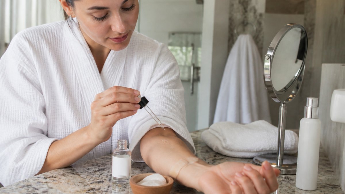A woman in a bathroom applying a small amount of clear gel from a dropper bottle to her forearm, with a patch test bandage and skincare products on the counter